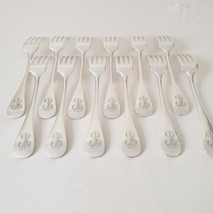 May include: A set of silver-coloured forks, each with a three-pronged head and a teardrop-shaped handle. The handles feature an engraved monogram design. The forks are arranged on a white surface, suggesting a formal dining setting.