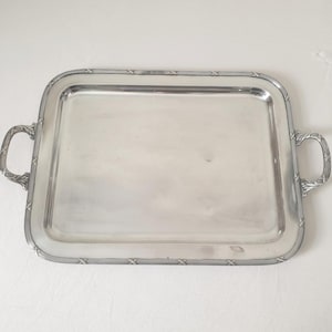 Può includere: Un vassoio da portata rettangolare argentato con due manici ornati. Il vassoio ha un bordo rialzato con un motivo decorativo attorno al bordo. Le maniglie sono curve e dettagliate, aggiungendo all'elegante design del vassoio. Il vassoio è probabilmente in metallo.