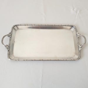 Könnte beinhalten: Ein rechteckiges silbernes Serviertablett mit verzierten Griffen und einem dekorativen Rand. Das Tablett besteht aus glänzendem, reflektierendem Metall und ist etwa 36 cm lang und 23 cm breit.