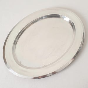 Può includere: Un vassoio da portata ovale argentato e lucido. Il vassoio ha un bordo rialzato e una superficie liscia e riflettente. Progettato per servire cibo o esporre oggetti, con un design classico.