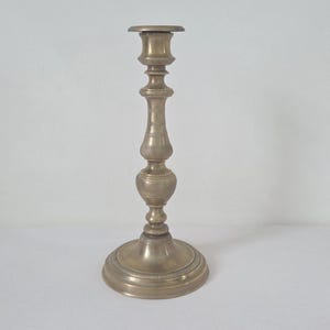 Könnte beinhalten: Ein Vintage-Kerzenhalter aus Messing mit rundem Sockel und aufwendigem Design. Der Kerzenhalter hat einen hohen, schlanken Stiel mit dekorativen Elementen und eine becherförmige Oberseite zur Aufnahme einer Kerze. Das Messing hat eine warme, gealterte Patina. Höhe: ca. 20 cm.