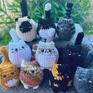 May include: A collection of handmade crochet cat plushies in various colors and patterns. The plushies feature detailed facial features, upright tails, and a soft, textured appearance. The cats are arranged together, showcasing the variety of designs.