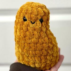 May include: A plush toy shaped like a peanut, made of yellow yarn with a fuzzy texture. The toy has black eyes and a smiling mouth.