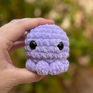 May include: A small, crocheted purple octopus with black eyes. The octopus is made with a soft, fuzzy yarn.