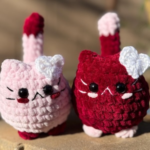 May include: Two crocheted cat plushies. One is light pink with a white heart and a red accent. The other is red with a white heart. Both have black eyes and a tail with a gradient of colors.