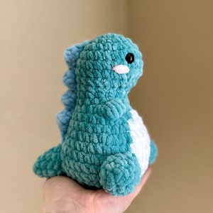 May include: A teal blue crocheted dinosaur plush toy with white accents. The dinosaur has a small, black eye and a white, stitched smile.