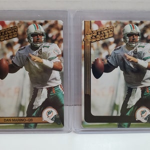May include: Two vintage trading cards of Dan Marino, a former American football quarterback, in a Miami Dolphins uniform. The cards are gold and black with the text "Action Packed" and "Dan Marino-QB".