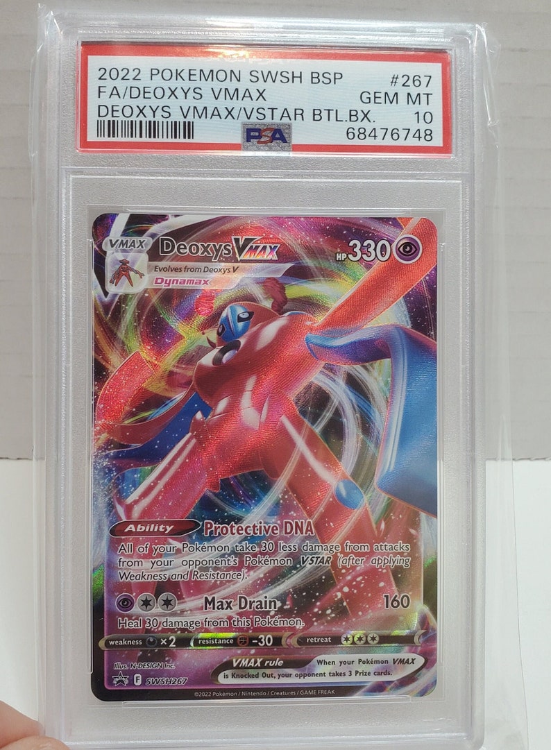 May include: A Deoxys VMAX Pokemon card from the Sword & Shield set. The card features a red and blue Deoxys VMAX with a purple background. The card is numbered 267 and has a Gem Mint condition rating.