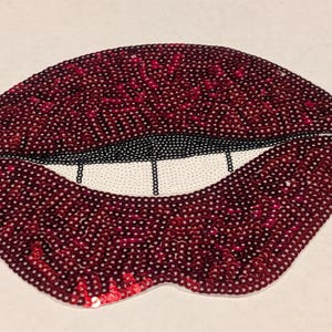 May include: A red sequin lips patch with a black outline and white teeth. The patch is made of sequins and is ideal for adding a touch of sparkle to any project.