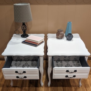 May include: Two white end tables with two drawers each. The drawers are open and lined with black and white floral patterned paper. One table has a lamp with a grey lampshade and a stack of books on top. The other table has a blue vase, a brown ceramic vase, and a black geometric vase.
