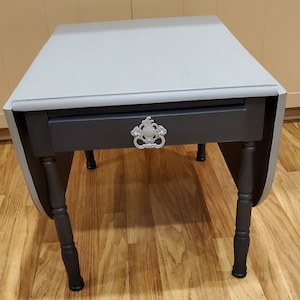 May include: A grey and black drop leaf table with a decorative silver knob. The table has four legs and a drawer.