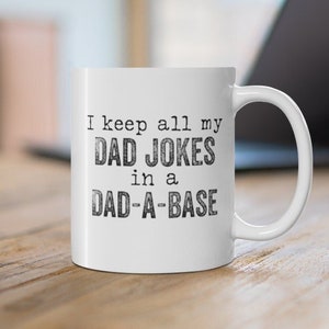 Op de afbeelding: Een witte keramische koffiemok met zwarte tekst die "I keep all my DAD JOKES in a DAD-A-BASE" zegt.