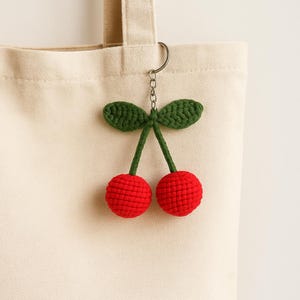 Puede incluir: Un llavero de cerezas rojas y verdes de ganchillo, colgado de un bolso de mano de lona. El llavero presenta dos cerezas rojas con tallos y hojas verdes, sujetas a un llavero plateado. La bolsa es de color beige claro.