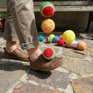 Puede incluir: Una persona con zapatos marrones y pantalones color caqui con varias bolas de ganchillo con forma de fruta en el aire. Las bolas están diseñadas para parecerse a una sandía, una naranja y un limón. Otras bolas coloridas están en el fondo.