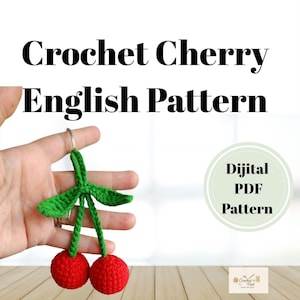 Puede incluir: Un llavero de cereza tejido a ganchillo con dos cerezas rojas y hojas verdes. El texto "Crochet Cherry English Pattern" aparece en la parte superior, junto con "Dijital PDF Pattern".