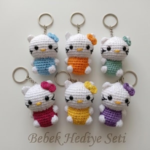 Peut inclure: Six porte-clés faits à la main en crochet en forme de chat de dessin animé, chacun avec une tenue et un nœud de couleur différente. Les porte-clés sont blancs avec des yeux noirs et un nez jaune. Les tenues sont bleues, orange, vertes, rouges, jaunes et violettes.