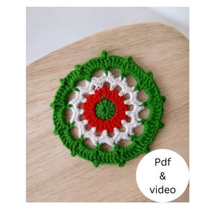 Könnte beinhalten: Ein handgefertigter Häkeluntersetzer mit kreisförmigem Design. Der Untersetzer hat eine rote Mitte, einen weißen Mittelring und einen grünen Außenring. Der Untersetzer wird auf einer Holzoberfläche präsentiert. Der Text auf dem Bild lautet "Pdf & video".