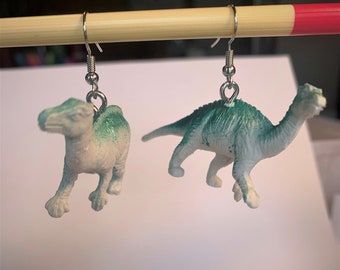 Könnte beinhalten: Ohrringe in Dinosaurierform. Jeder Ohrring zeigt eine kleine, weiße Dinosaurierfigur mit grünen Akzenten, die an einem silberfarbenen Haken befestigt ist. Die Ohrringe sind so konzipiert, dass sie baumeln und die einzigartigen Dinosaurierfiguren zeigen.
