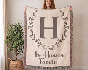 May include: A cream-colored woven throw blanket featuring a large dark gray letter H centered within a decorative leaf wreath. Below the wreath, the text reads EST. 2012 and The Hanson Family. The blanket has fringed edges with multicolored threads.