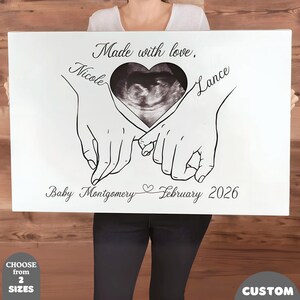 May include: White canvas art featuring a sonogram image within a heart shape, held by two hands. The names "Nicole" and "Lance" are written on the canvas, along with "Baby Montgomery February 2026" and "Made with love."