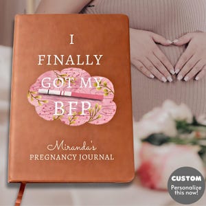 Puede incluir: Un diario de embarazo de cuero marrón con el texto "I FINALLY GOT MY BFP". El diario presenta un diseño de nube rosa con una ilustración de prueba de embarazo positiva y las palabras "Miranda's PREGNANCY JOURNAL" debajo.