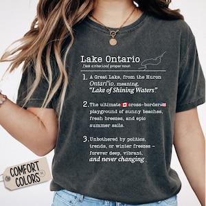 May include: A charcoal grey short-sleeve t-shirt featuring white text that defines Lake Ontario with a dictionary-style entry, including a map outline, the phrase Lake of Shining Waters, and small Canadian and American flag icons.