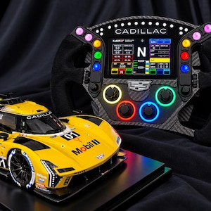 May include: A yellow Cadillac race car model with number 01 and Mobil 1 branding, positioned next to a black carbon fibre racing steering wheel featuring a digital display, illuminated buttons, and the Cadillac logo in the centre.