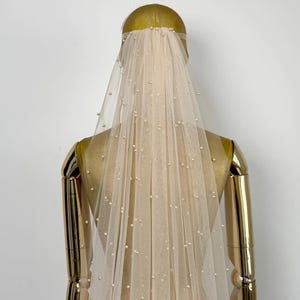 May include: A white tulle bridal veil with pearl embellishments. The veil is draped over a mannequin head.