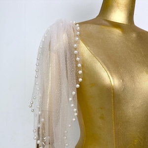 Puede incluir: Una manga transparente de color blanco roto con un delicado estampado de lunares y un ribete de pequeñas perlas redondas. La manga está drapeada sobre un maniquí dorado, creando un aspecto suave y elegante. El diseño es adecuado para ropa formal.