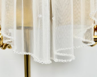 May include: A white bridal veil with a beaded edge. The veil is displayed on a gold mannequin.