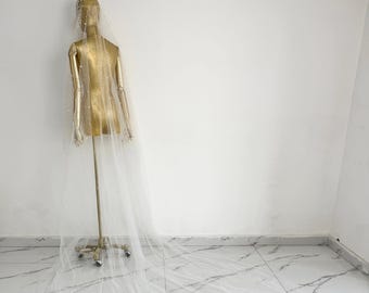 May include: A long, white bridal veil with a beaded trim. The veil is draped over a mannequin and is laying on a white marble floor.