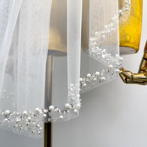 May include: White bridal veil with a pearl and rhinestone trim. The veil is made of soft tulle and is perfect for a wedding or special occasion.