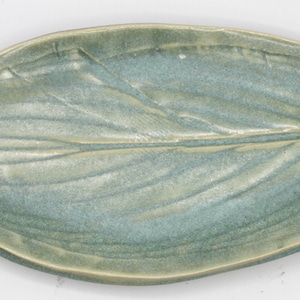 May include: A ceramic leaf-shaped dish with a light blue glaze. The dish has a detailed vein pattern and a slightly raised edge.