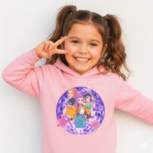 May include: A light pink hooded sweatshirt featuring a graphic of four cartoon characters and a blue cat in front of a purple disco ball. The sweatshirt has long sleeves and a drawstring hood. The girl in the image is smiling and giving a peace sign.