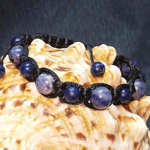 May include: A blue gemstone bracelet with black cord and knots. The bracelet is on a seashell.