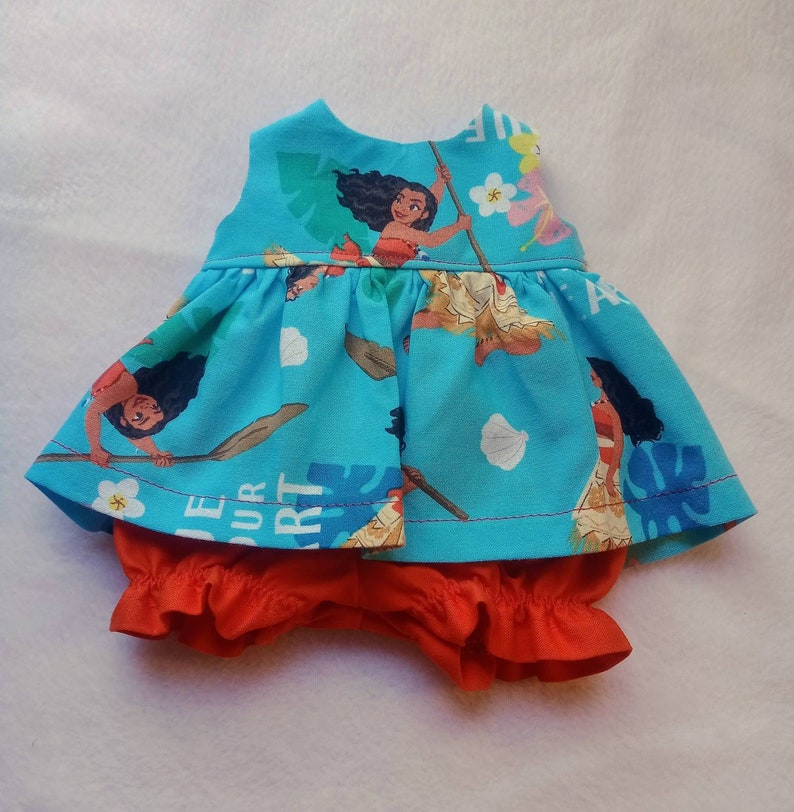 May include: A blue and red doll's dress with a sleeveless bodice and a ruffled skirt. The dress features a print of Moana from the Disney film Moana. The dress is paired with red ruffled bloomers.