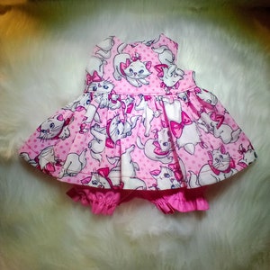 May include: A pink and white baby dress with a polka dot pattern and a print of cartoon cats. The dress has a sleeveless bodice and a ruffled skirt. The dress is paired with a pink ruffled diaper cover.