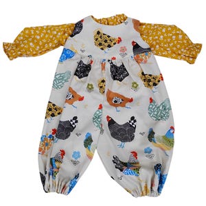 May include: A baby's jumpsuit with a white background and a pattern of colorful chickens. The long sleeves are mustard yellow with a small floral print. The jumpsuit has a ruffled neckline and cuffs.