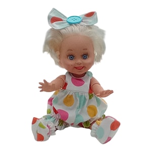 May include: A vintage doll with white hair, blue eyes, and a smiling expression. The doll wears a polka-dot dress, matching pants, and a bow with a blue button. The doll is sitting with its arms outstretched.