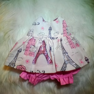 May include: A white and pink baby dress with a Paris theme. The dress features a print of the Eiffel Tower, buildings, and clouds. The dress has a pink ruffle trim and a matching pink diaper cover.