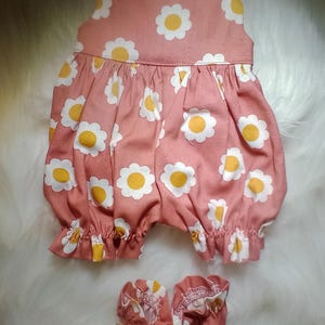 May include: A pink baby doll outfit with a daisy print and matching booties. The outfit has a gathered skirt and ruffled trim.