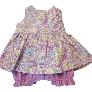 May include: A sleeveless floral print top and matching bloomers set. The top features a white background with a colorful floral pattern in shades of purple, yellow, and blue. The bloomers are a solid light purple color.
