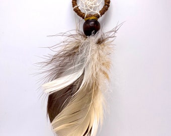 May include: A small brown dreamcatcher with white netting and a brown wooden bead hanging from a brown cord. The dreamcatcher has a single brown feather attached to the bottom.