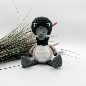 May include: A handmade crochet duckling toy. The duckling has a black head, a gray beak, and large, round eyes. Its body is white, gray, and brown. The toy is sitting on a white surface, with some decorative grass in the background.