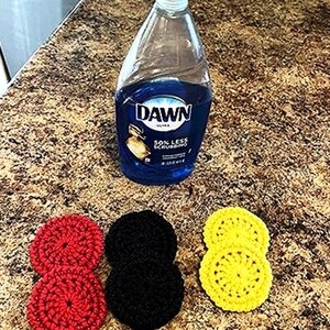 May include: Three crocheted dishcloths in red, black, and yellow. The dishcloths are stacked on top of each other. A bottle of Dawn dish soap is in the background.