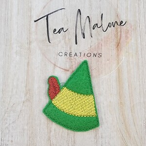 May include: A green felt elf hat with a gold band and a red ear. The hat is on a light wood grain surface. The text "Tea Malone Creations" is in the background.