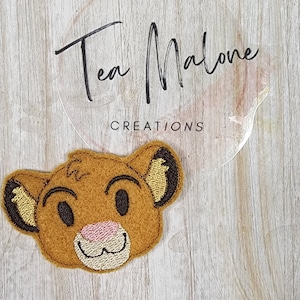 Puede incluir: Una aplicación de fieltro de Simba de El Rey León. La aplicación es marrón con la nariz rosa y los ojos negros. Es un diseño lindo y caprichoso.