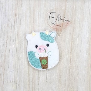 May include: White cow with blue spots holding a brown and white coffee cup with a green lid and a straw. The cow is embroidered on a white fabric.