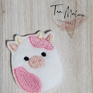 May include: A white felt cow applique with pink spots and a gold embroidered nose. The cow has black eyes and a pink mouth.