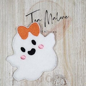 May include: A white felt ghost with a black smiling face and pink cheeks. The ghost has an orange felt bow on top of its head.